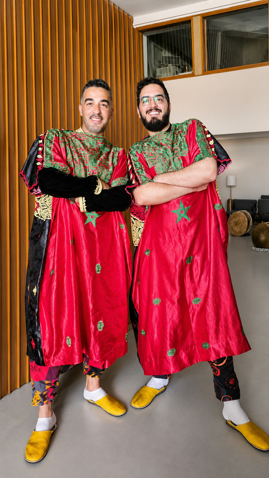 Two smiling band members in red kaftans with green embroidered yokes stand arms crossed before a slatted wood wall, drums resting behind them