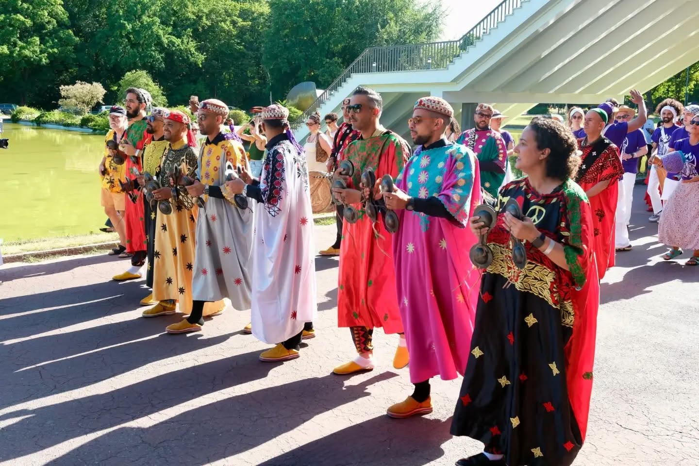 A line of musicians in brightly coloured robes plays qraqeb while walking beside a lake in the sunshine